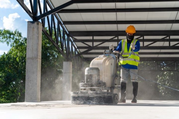 Worker in safety gear grinding a concrete floor under a metal roof; dust visible.