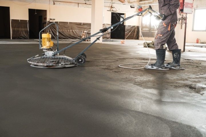 Worker smoothing concrete floor with power grinder in unfinished building.