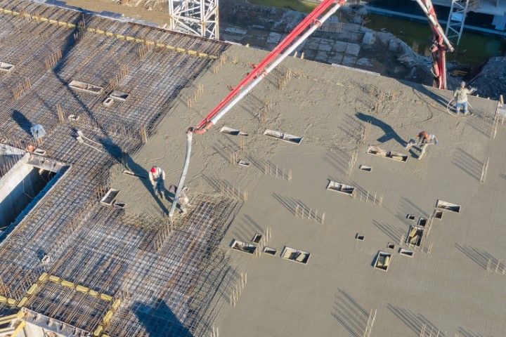 Construction workers pouring concrete on a building's roof, using a boom pump. Rebar is visible.