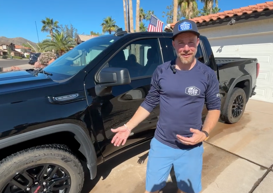 A person in a long-sleeve shirt and cap gestures toward a black pickup truck parked in a sunny residential driveway.
