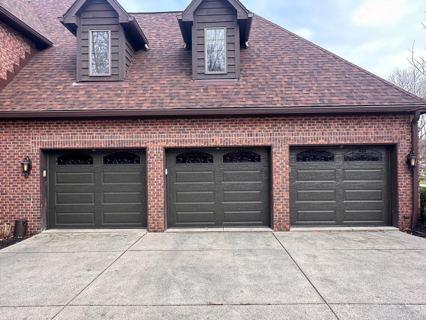 Three dark brown garage doors on a brick house with two small windows above.