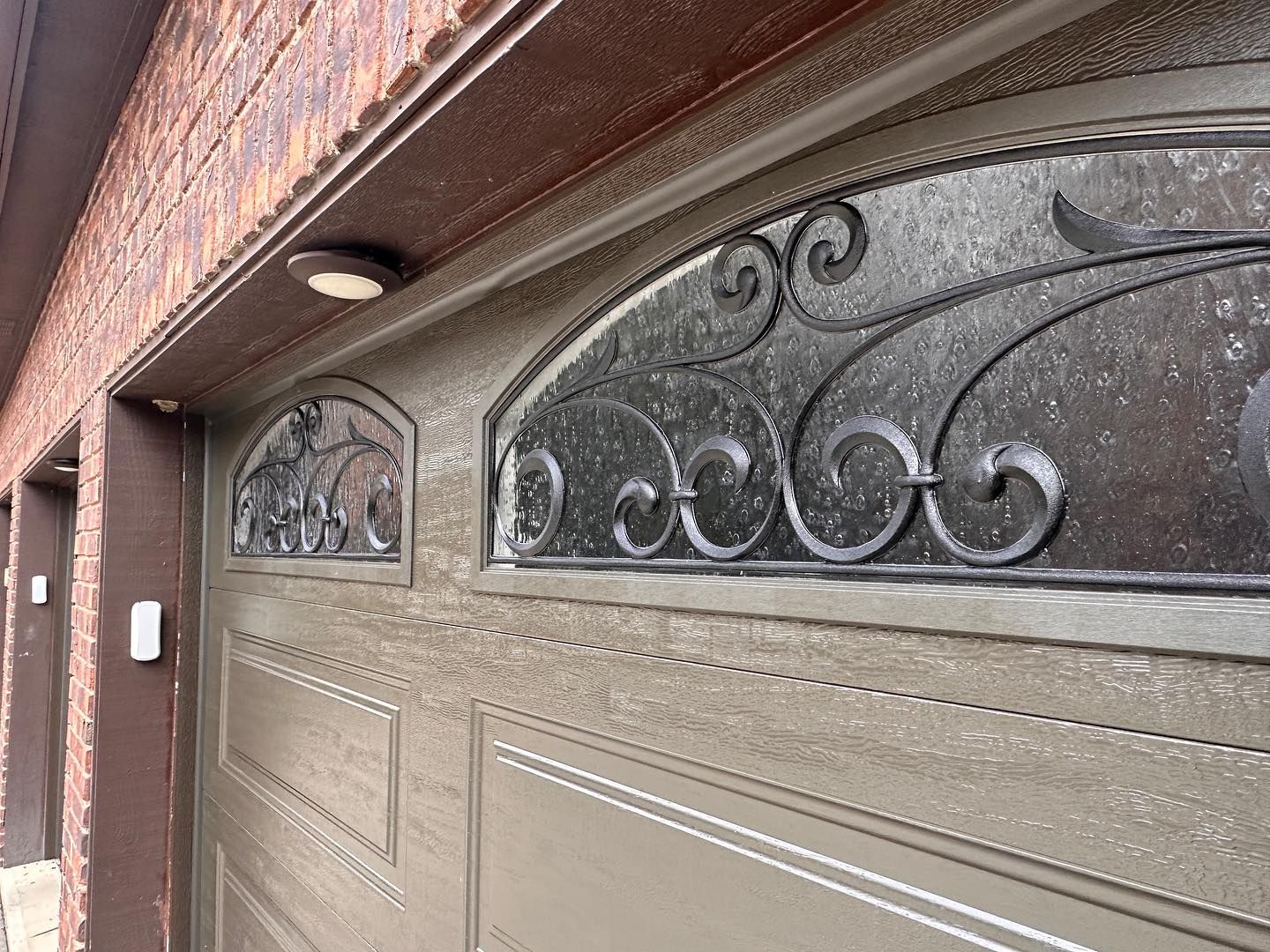 Tan garage door with decorative wrought iron design and glass panels. Brown trim.