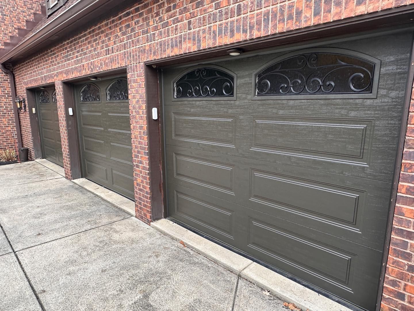 Three dark green garage doors with decorative top panels set in a brick building.