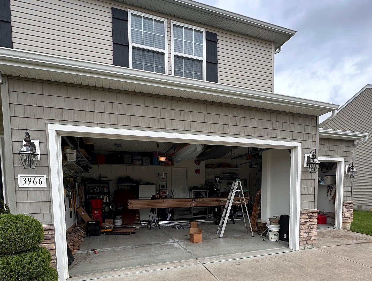 Two-car garage open with construction materials visible. Gray siding, white trim, and a ladder are inside.