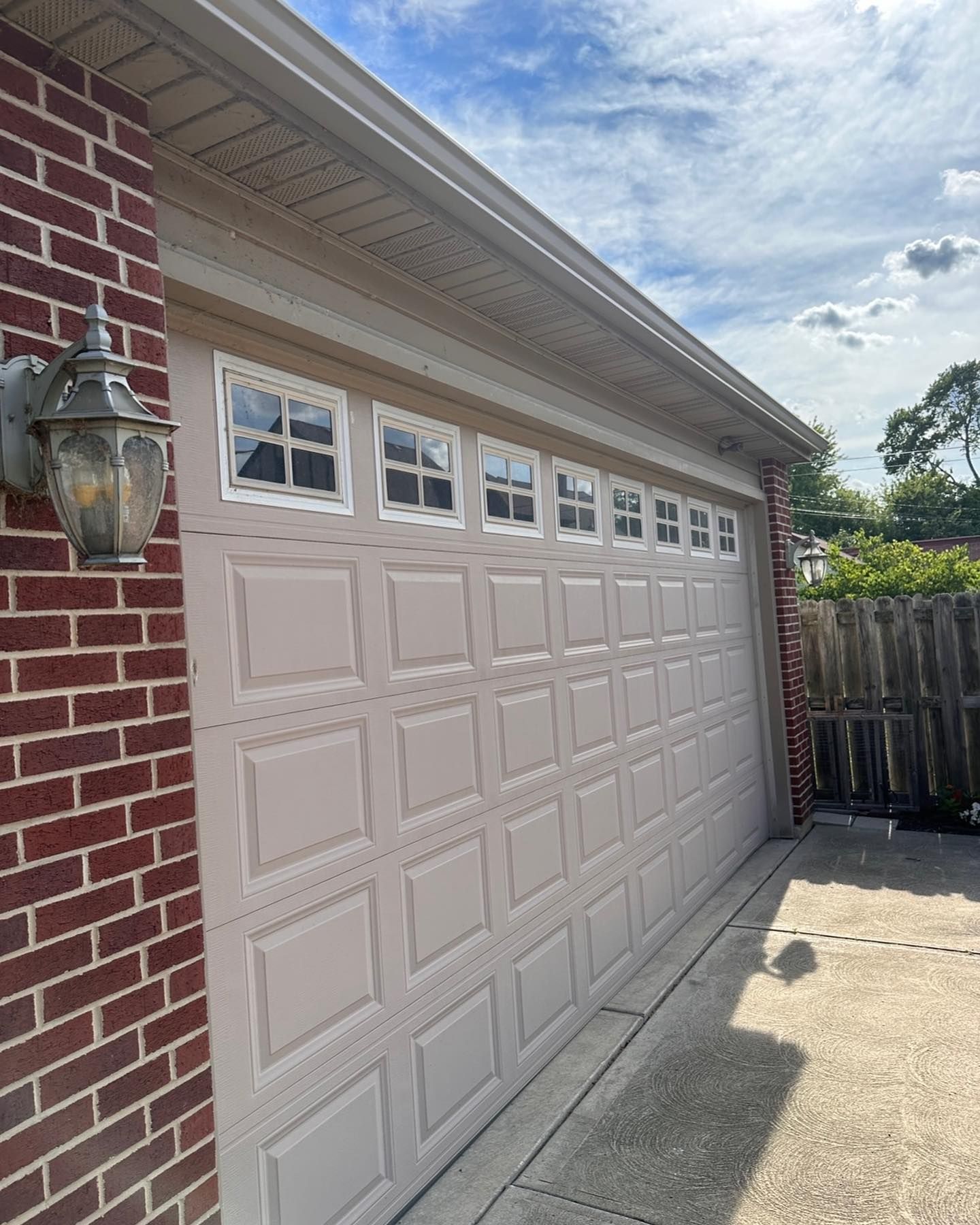 Tan garage door with square windows, brick wall, and a driveway. Blue sky with clouds.