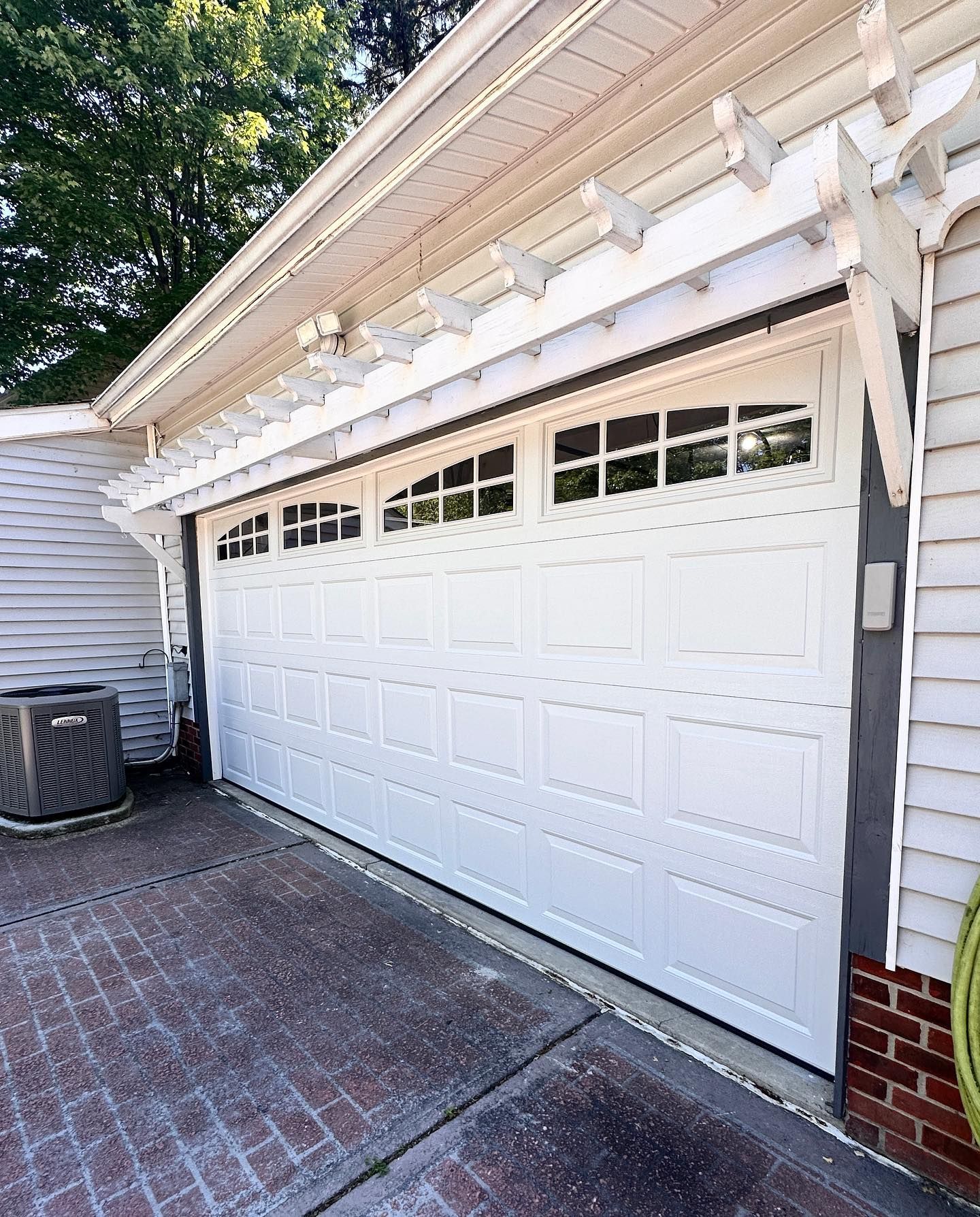 White garage door with windows, beneath a pergola. Brick driveway and gray siding.