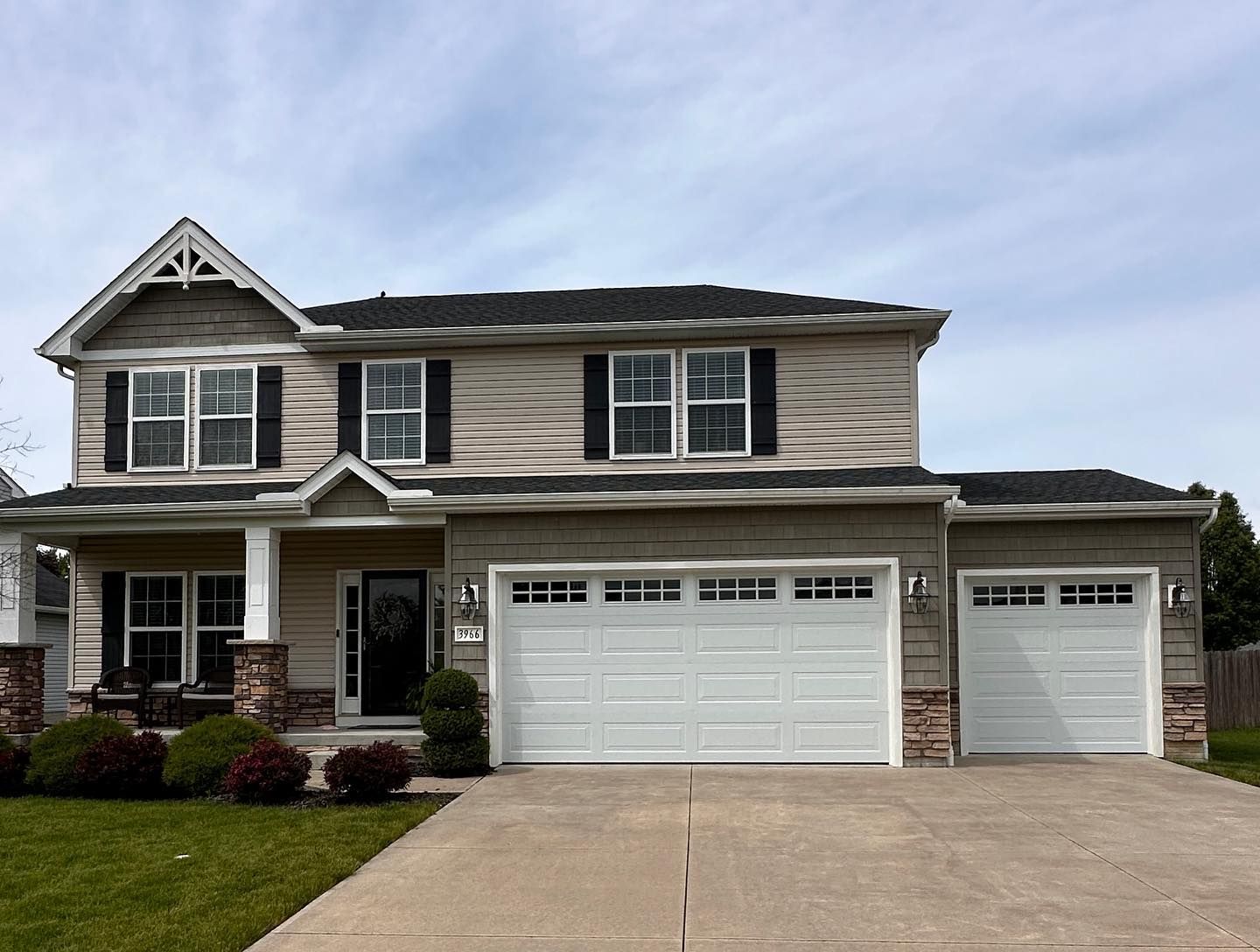 Two-story house with a garage, beige siding, black shutters, and a concrete driveway under a cloudy sky.