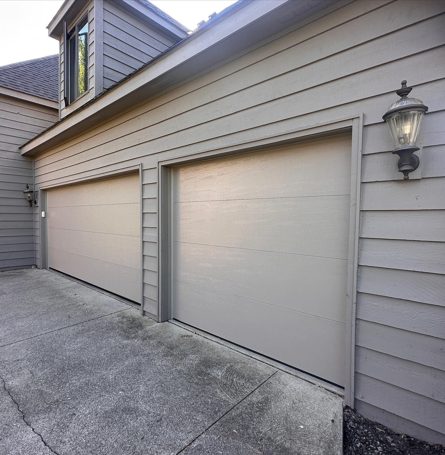 Two beige garage doors on a gray house with gray siding. A wall lamp is mounted to the right.
