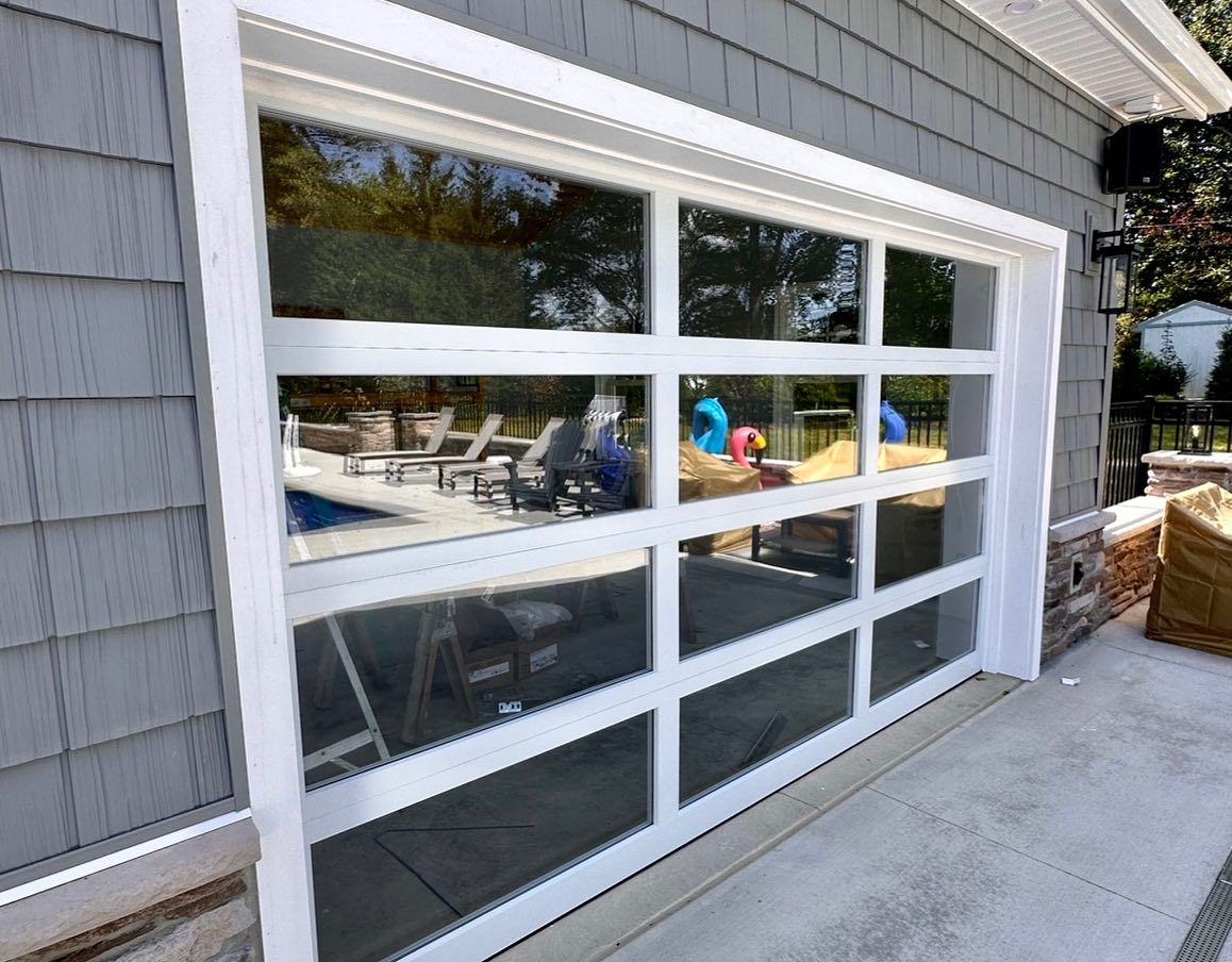 White-framed, glass garage door on a gray shingled building, reflecting a pool area.