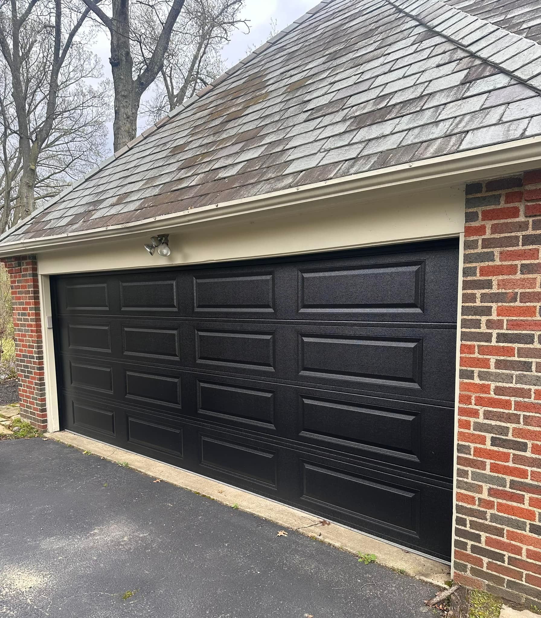 Black garage door on a brick building, with a driveway and a gray roof.