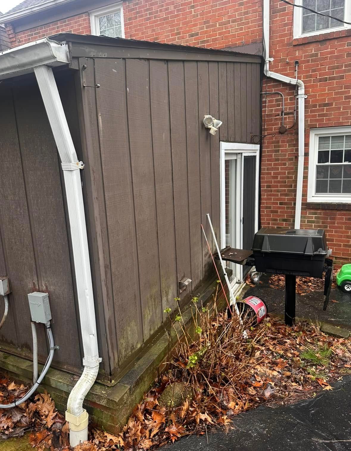 Brown shed attached to a brick building with a grill outside and overgrown brush.