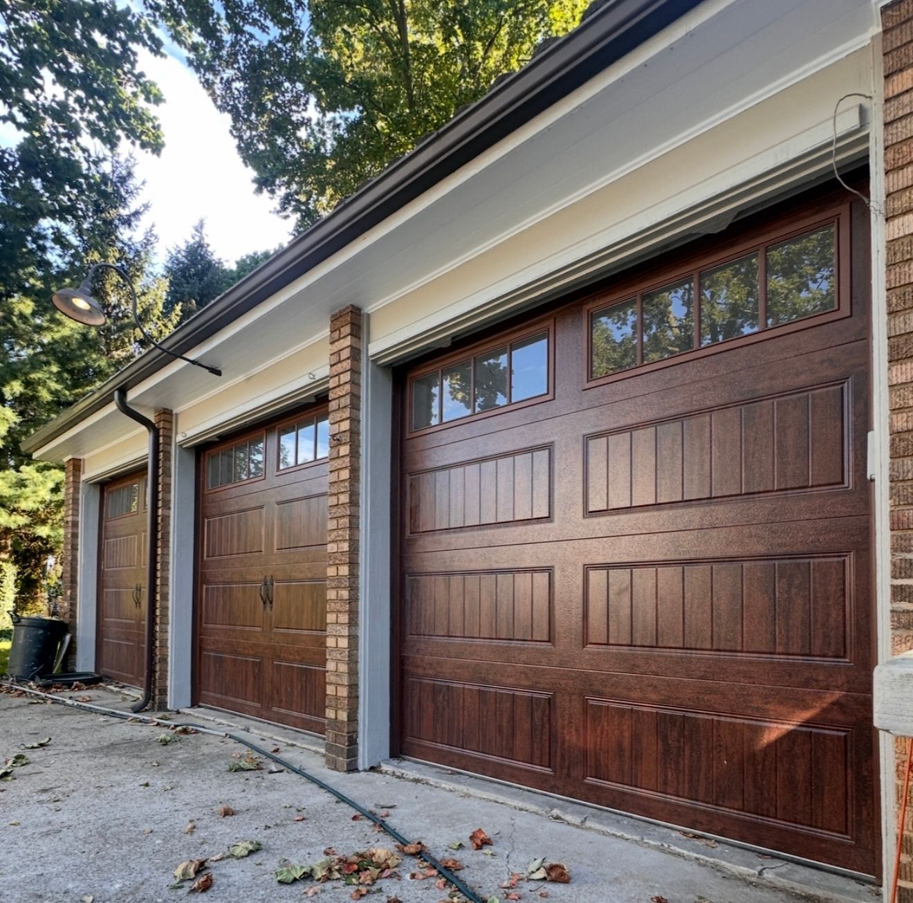 Three dark brown garage doors with glass windows. Brick facade, light trim, trees in background.