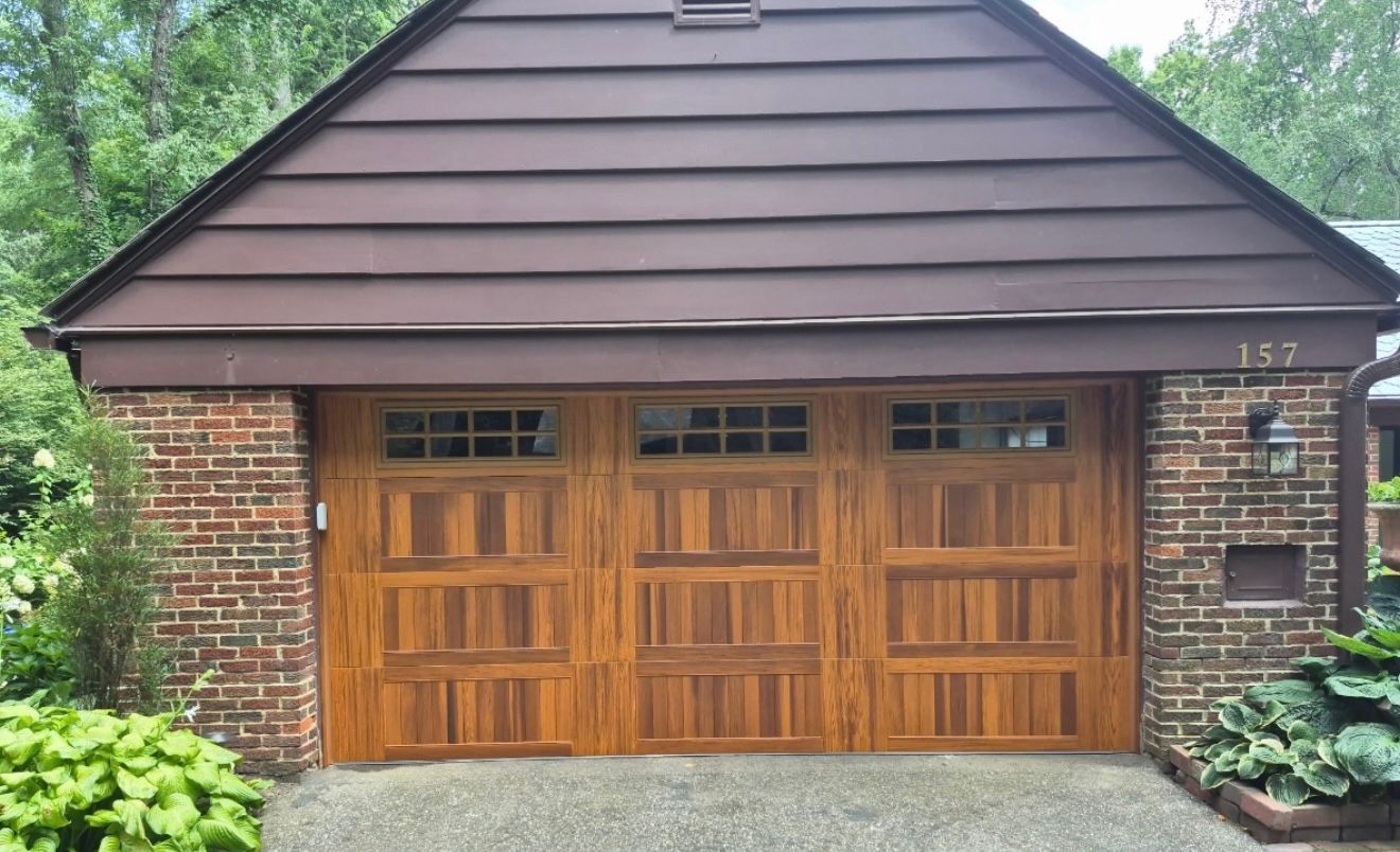 Brown wooden garage doors, brick columns, brown siding, and a green yard.