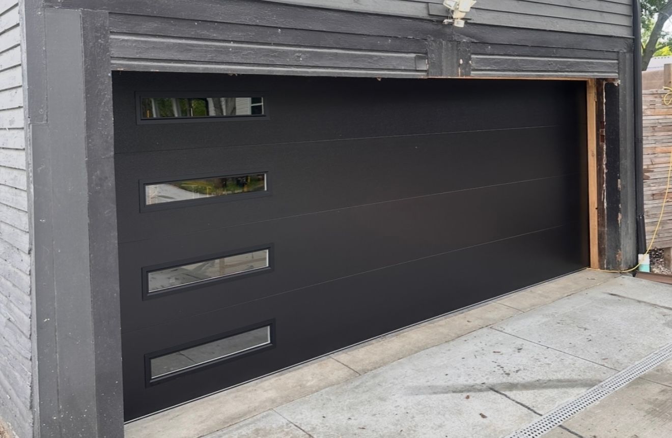 Black modern garage door with rectangular glass windows, set in gray building.