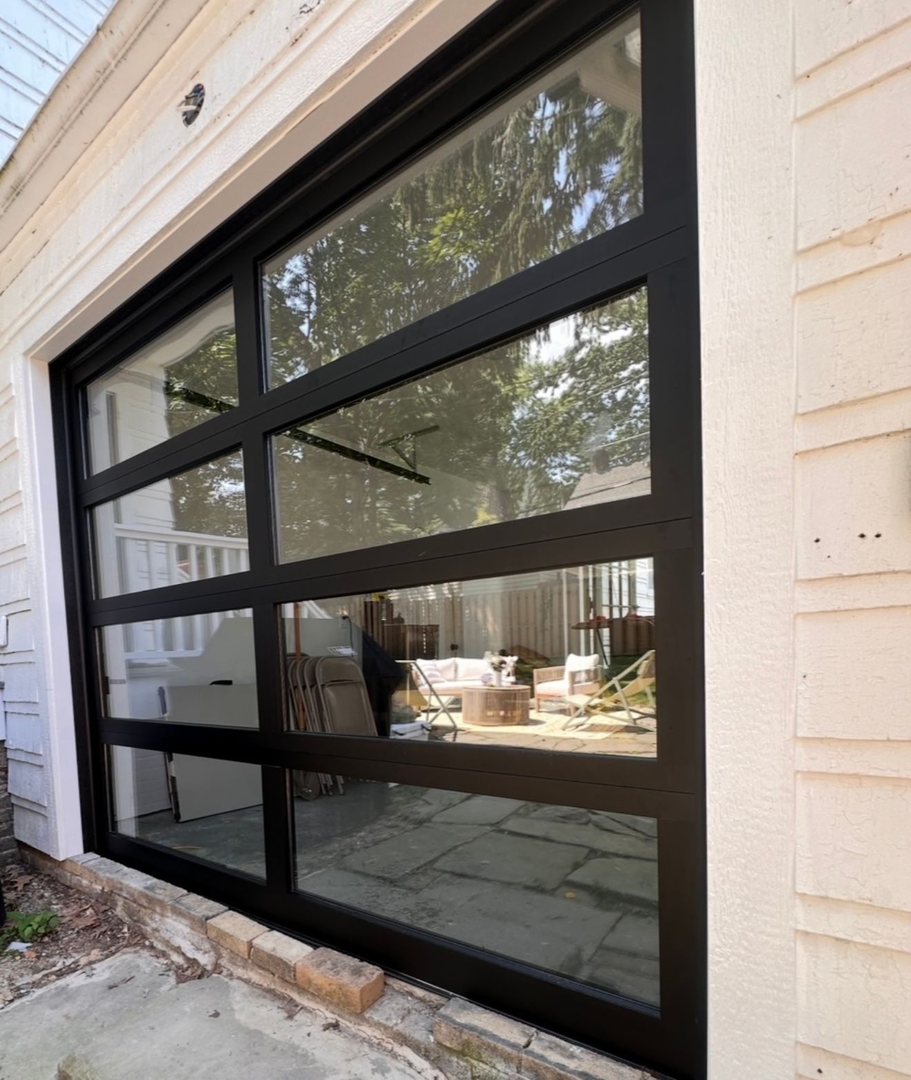 Black-framed glass garage door reflecting backyard patio, trees, and a person; attached to a white building.
