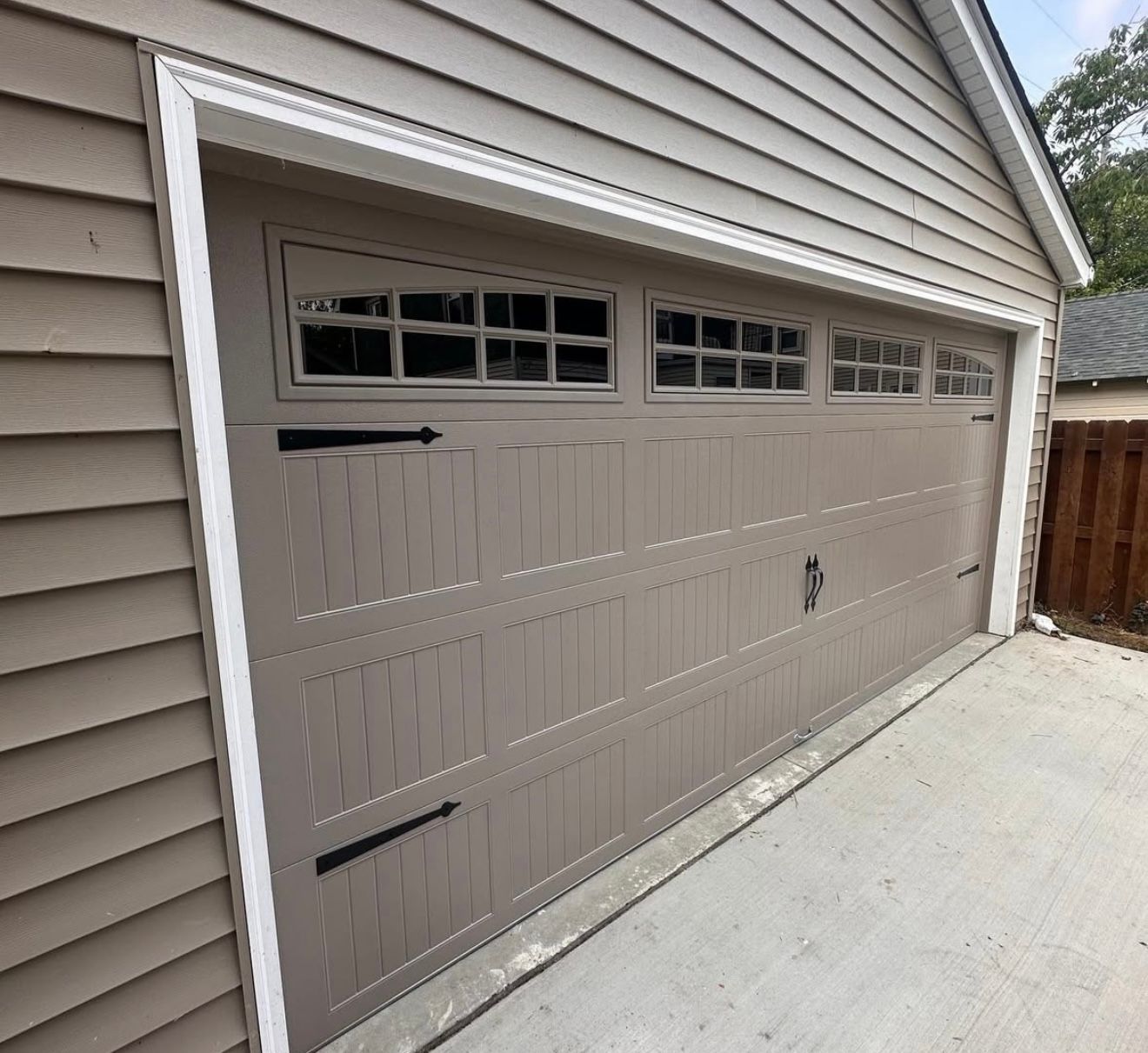 Tan garage door with decorative hardware and upper windows, set in a beige-sided building.