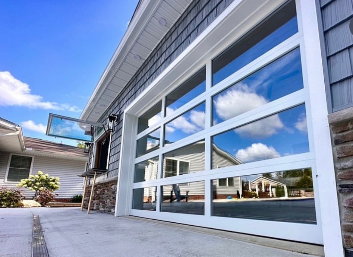White-framed glass garage door reflects sky and nearby houses; set in an overcast sky with gray siding.
