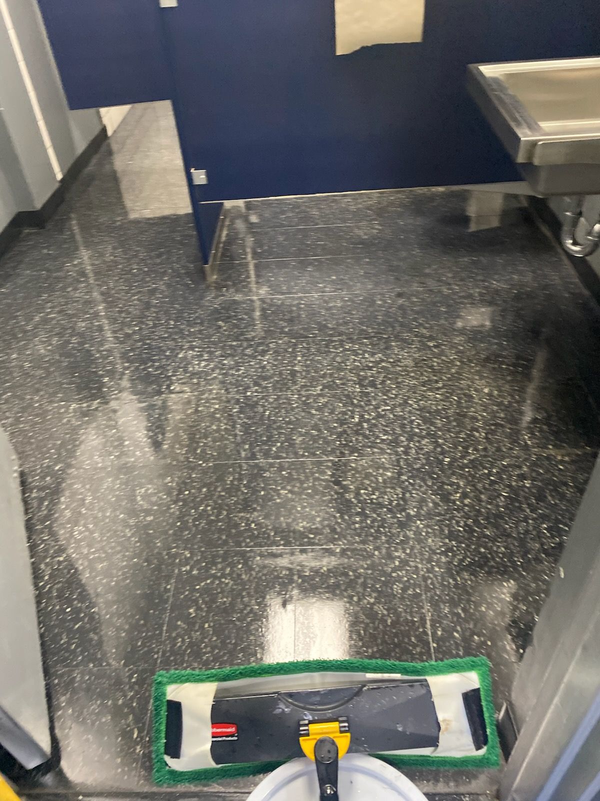 Medical room floor with a green cleaning mop in the foreground and a metal sink in the corner.