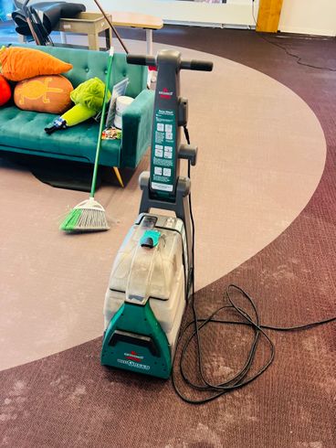 Upright carpet cleaner on a rug in a living room, with a cord and colorful cushions nearby