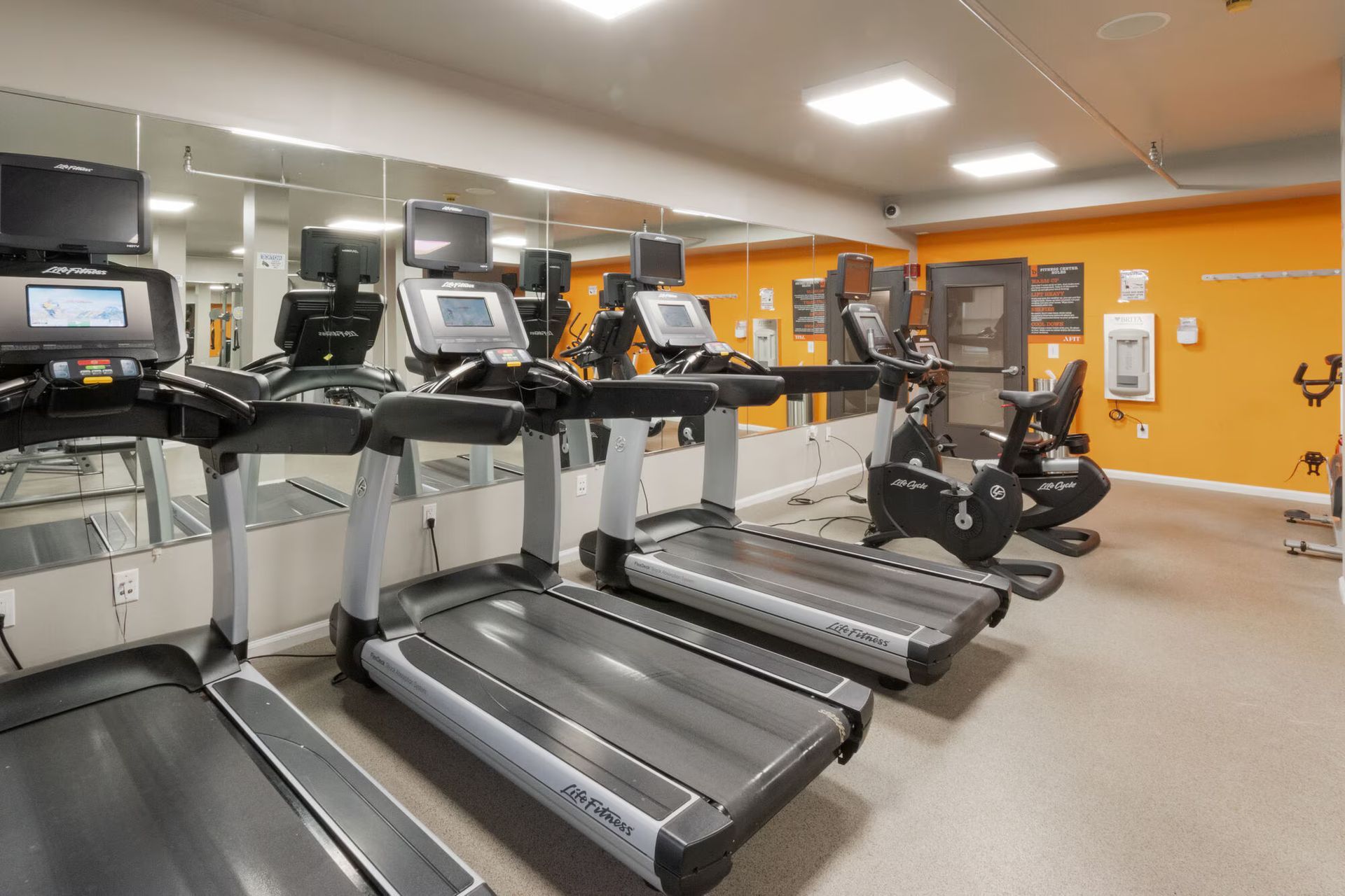 Gym with treadmills, stationary bikes, mirrors, and orange wall. Our pet-friendly apartments in New Brunswick, NJ offer convenient amenities, like a clubhouse, fitness center, and package lockers. Visit us today for a tour!
