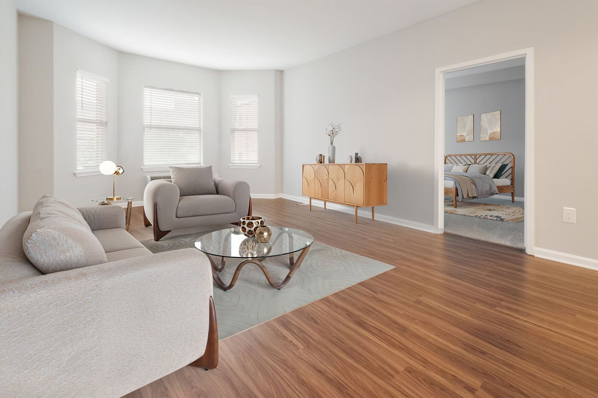 Living room with light gray sofa, chair, wooden cabinet, and view to a bedroom. Contact us to take a tour of our studio, 1, and 2-bedroom apartments for rent in New Brunswick, NJ today!
