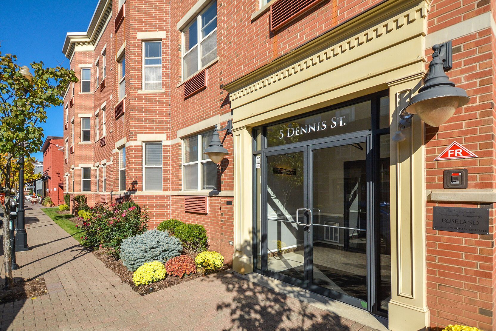 Brick apartment building entrance, sidewalk with flowers, sunny day. 