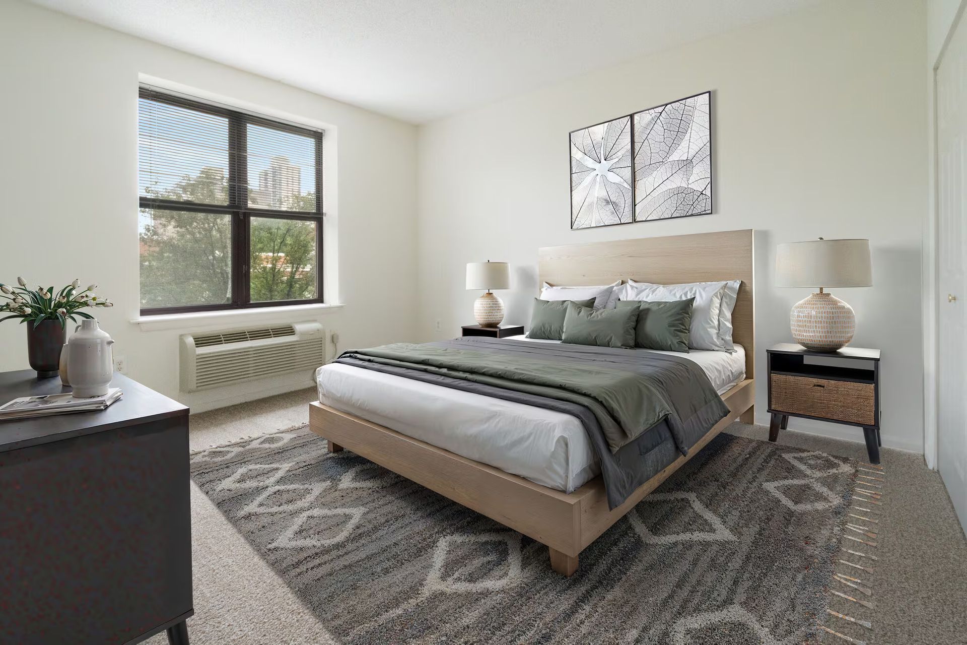 Bedroom with bed, nightstands, rug, window, and artwork. Neutral color scheme, natural light. Make The Brunswick your home today! Contact us to schedule a tour of our studio, 1, and 2-bedroom New Brunswick apartments for rent. 