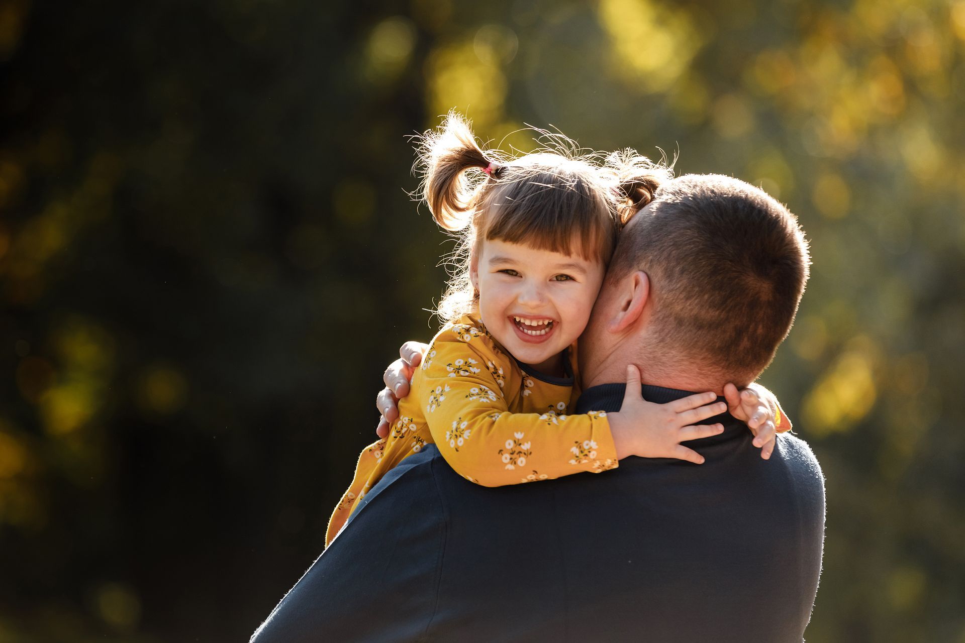 Lächelndes Kind mit Zöpfen umarmt eine Person von hinten, im Freien vor einem verschwommenen grünen Hintergrund.