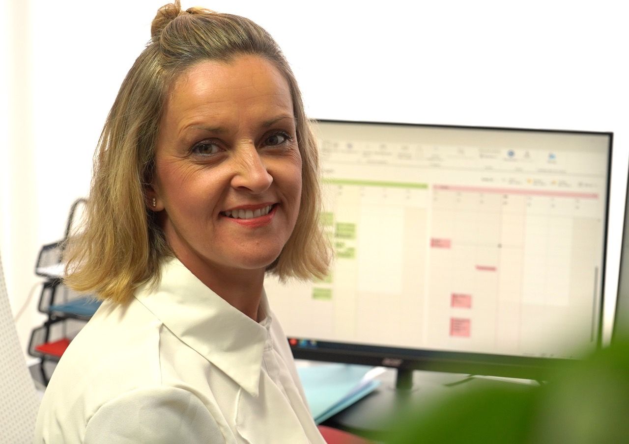 Frau lächelt in die Kamera, während sie an einem Computer arbeitet, auf dem ein Zeitplan sichtbar ist.