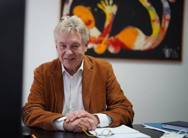 Mann in brauner Jacke sitzt lächelnd am Schreibtisch. Abstraktes Gemälde an der Wand dahinter.