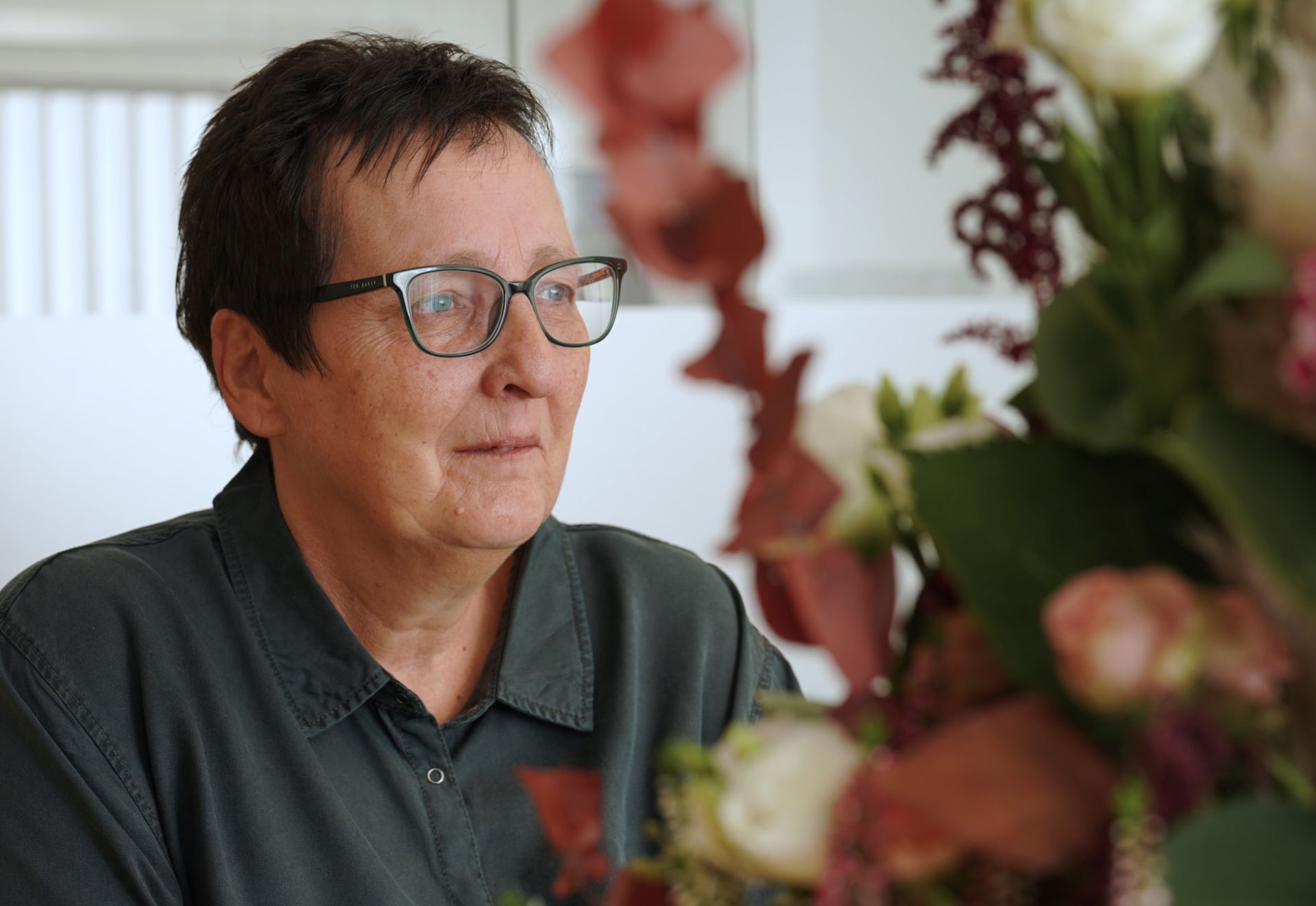 Frau mit Brille lächelt neben einem Blumenarrangement, sitzt im Haus.