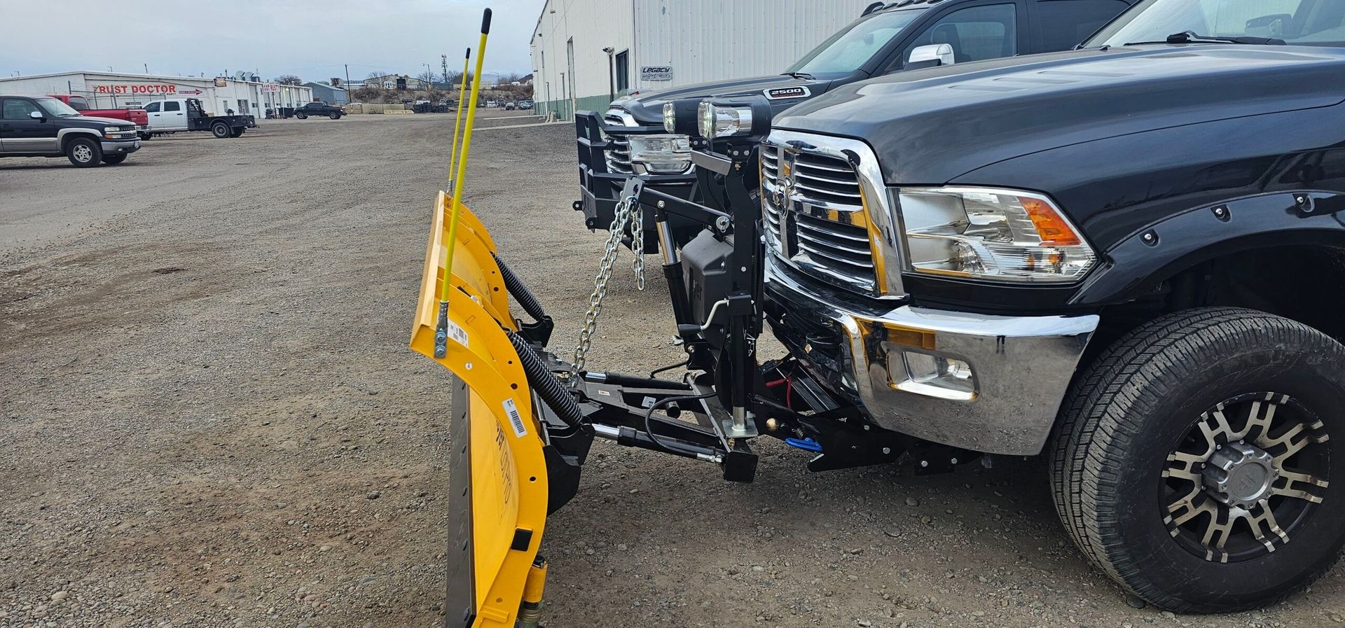 A black truck with a yellow snow plow attached to it is parked in a gravel lot.