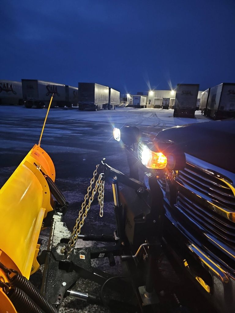 A snow plow is attached to the front of a truck in a parking lot at night.