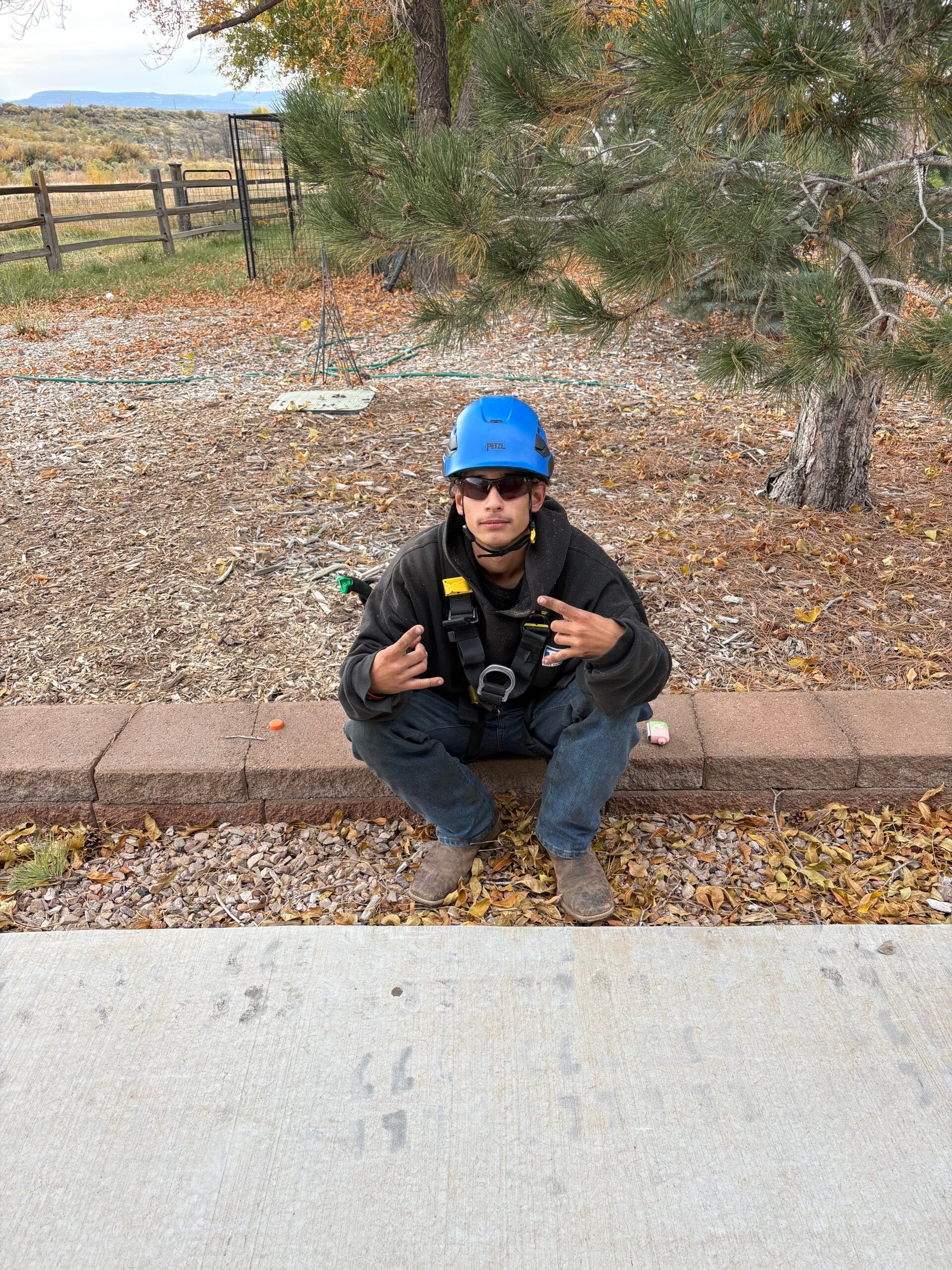 A man wearing a hard hat and sunglasses is squatting down on the sidewalk.