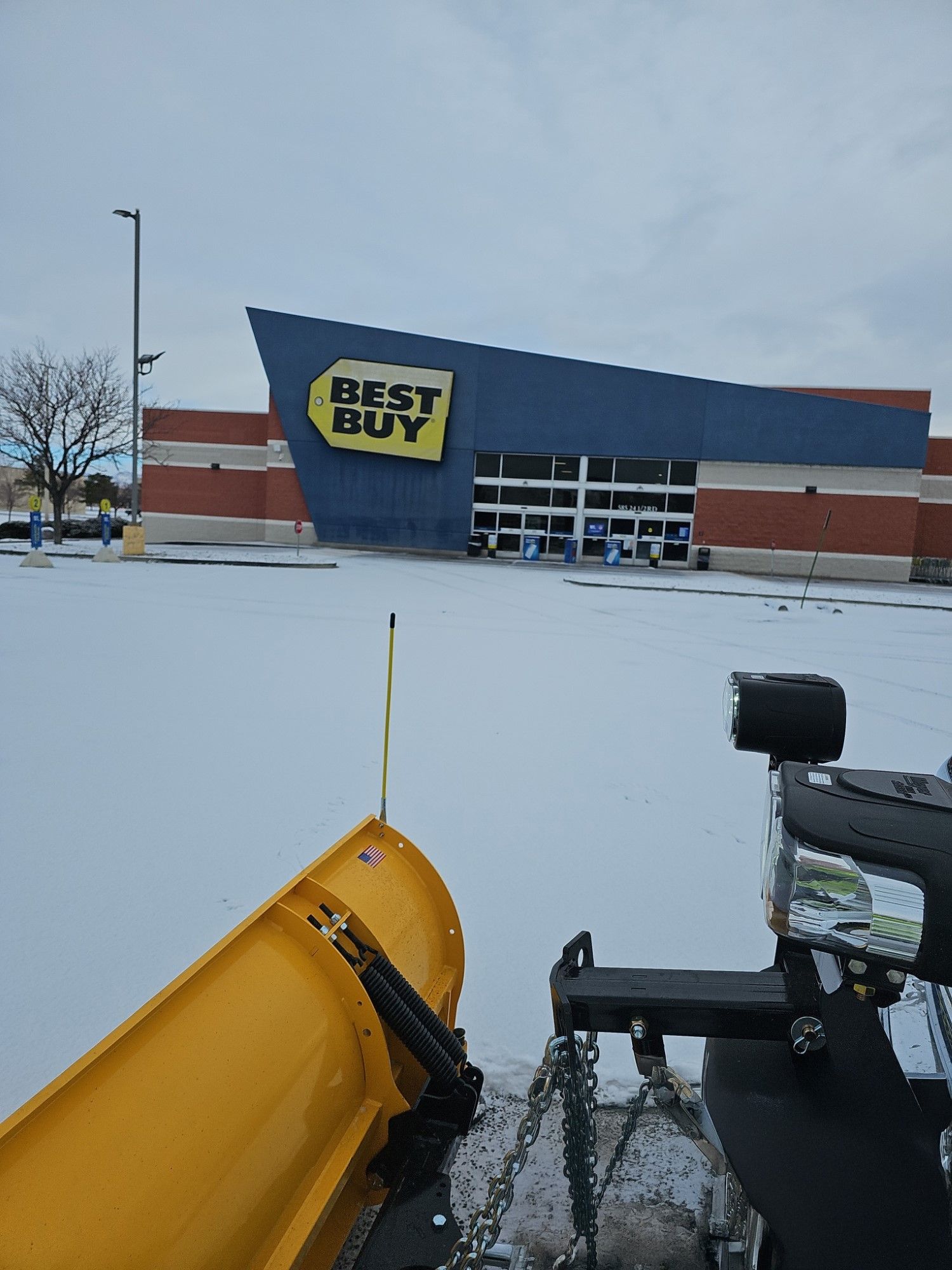 A yellow plow is plowing snow in front of a best buy store