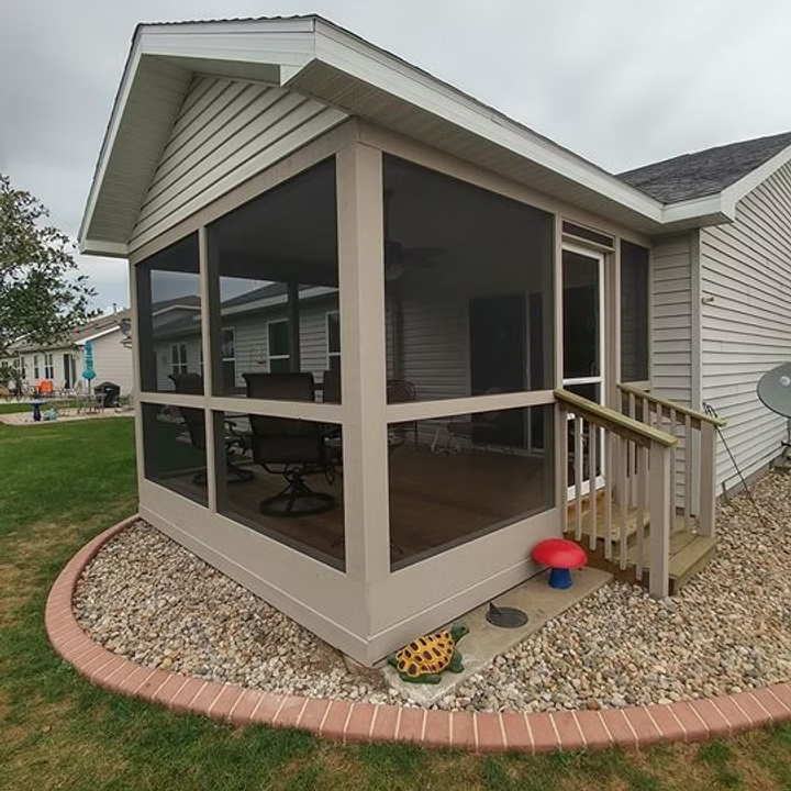 Enclosed screened porch with beige siding, dark screens, and a small ramp over a rock border.