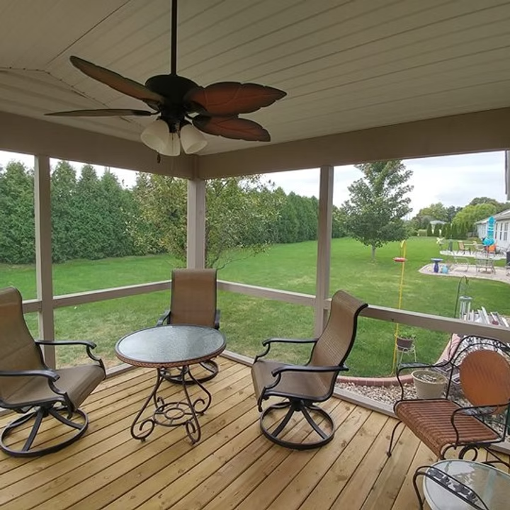 Covered wooden porch with rocking chairs, round table, ceiling fan, and view of a green backyard and pool