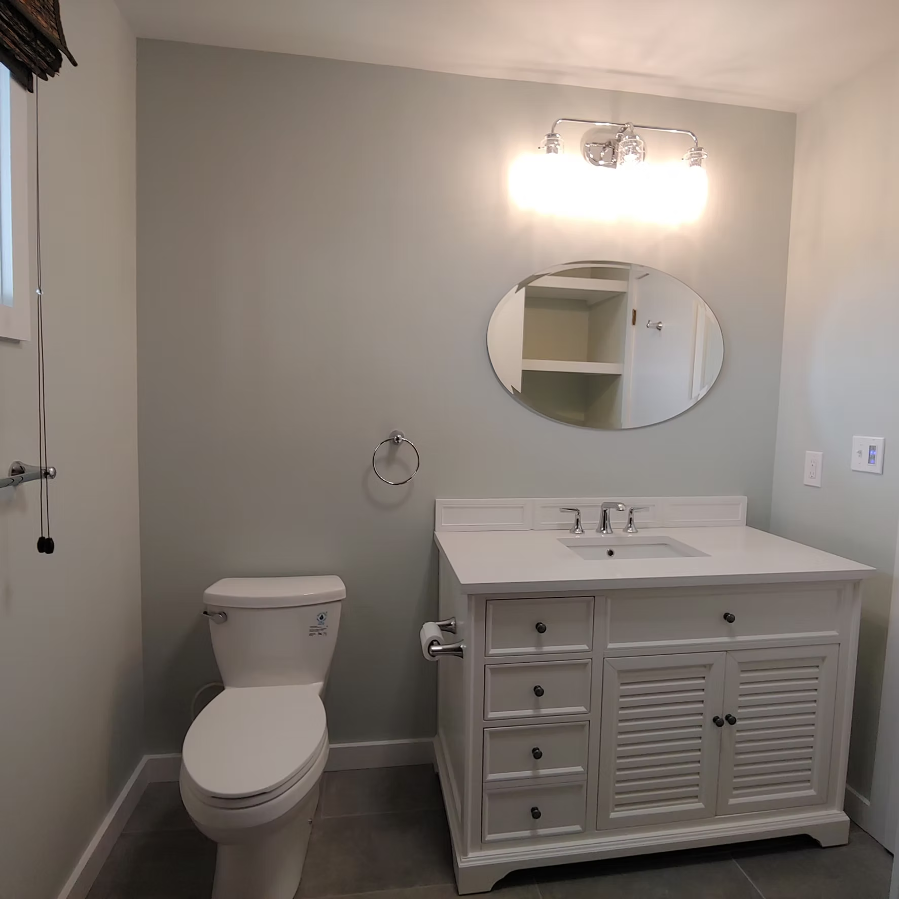 Small bathroom with white vanity, toilet, round mirror, and wall light on gray walls.