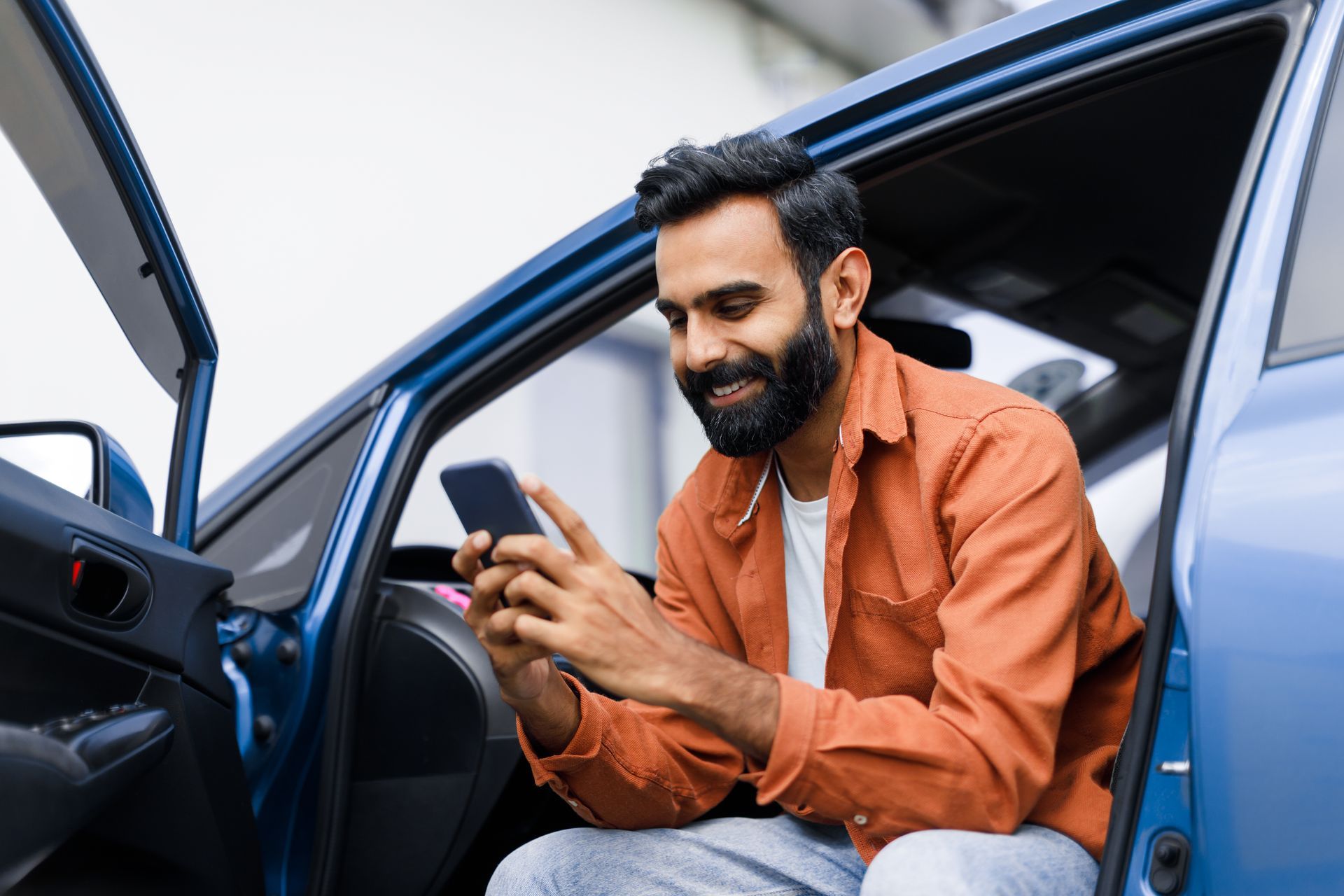 Man sitting in a blue car doorway, smiling at his phone, wearing an orange shirt.