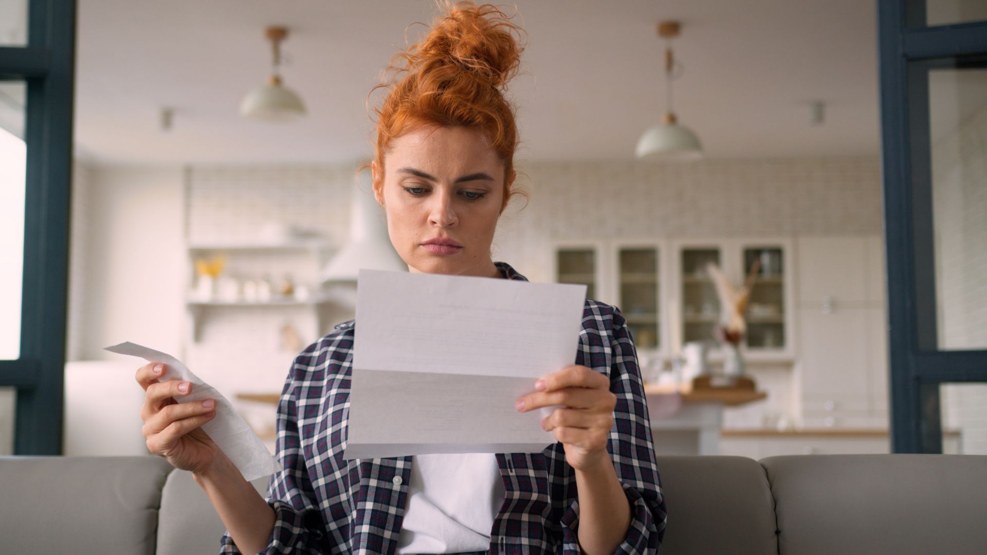 Person in a plaid shirt reading a letter on a couch in a bright living room
