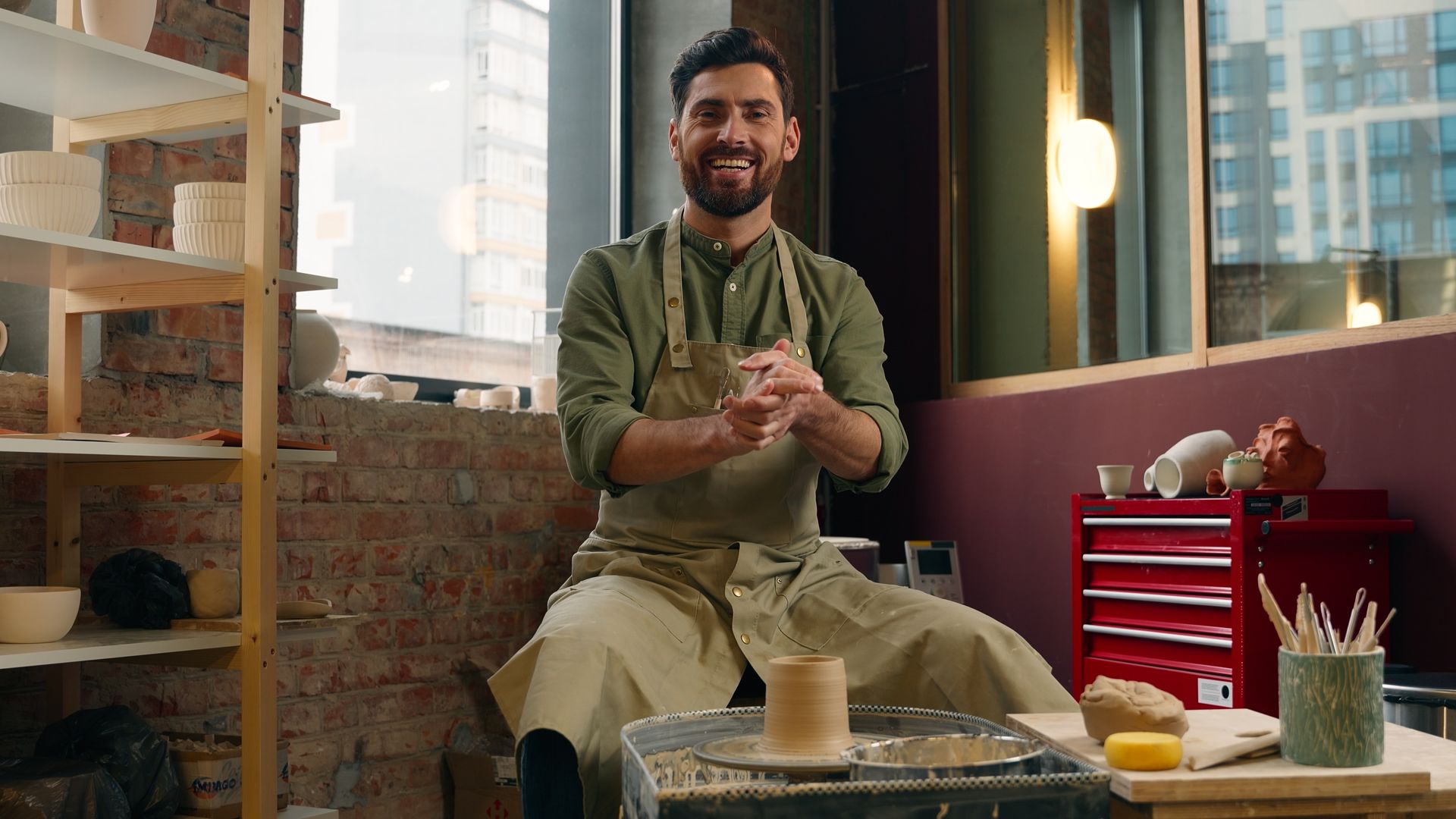 Man sitting on a stool in a cozy loft studio, smiling and clapping beside a red tool chest and potted plants