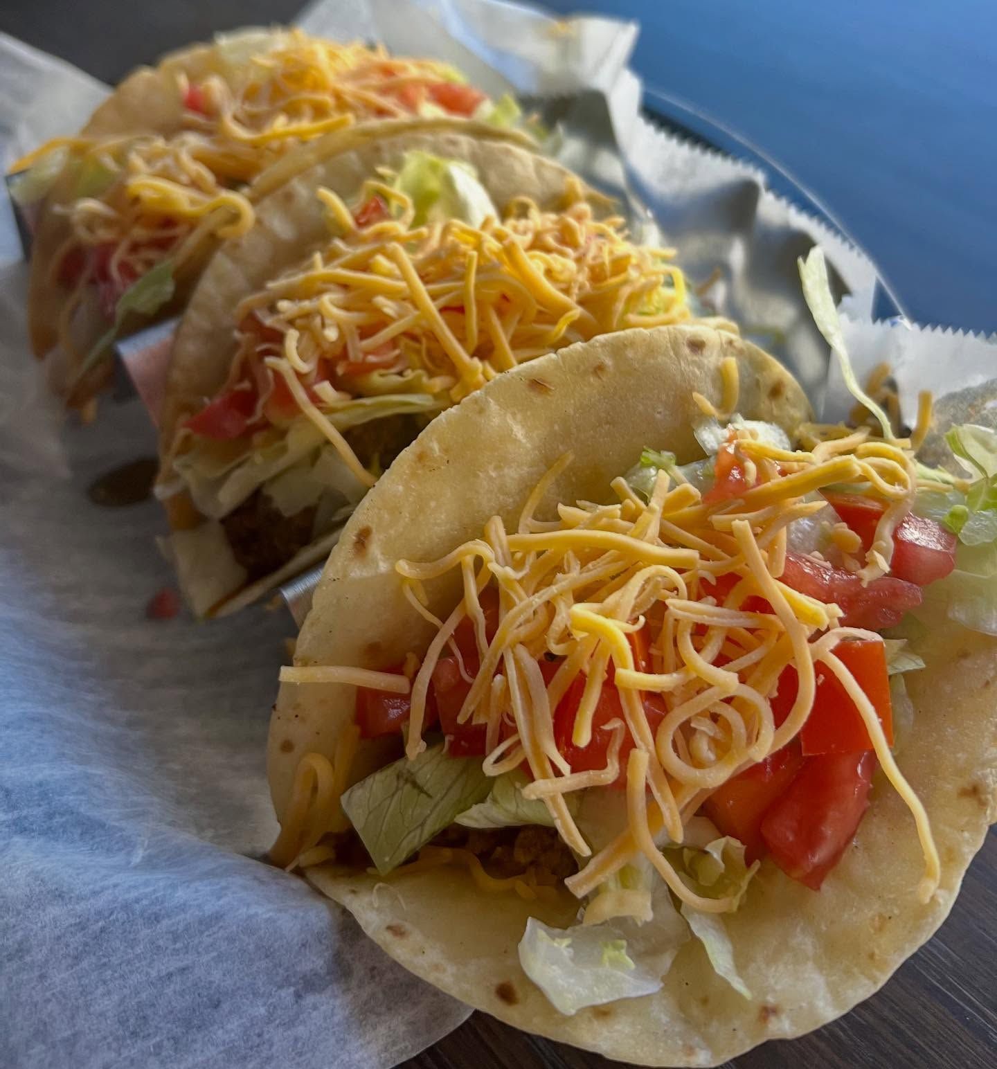 Four tacos in a basket, with shredded cheese, lettuce, and tomatoes.