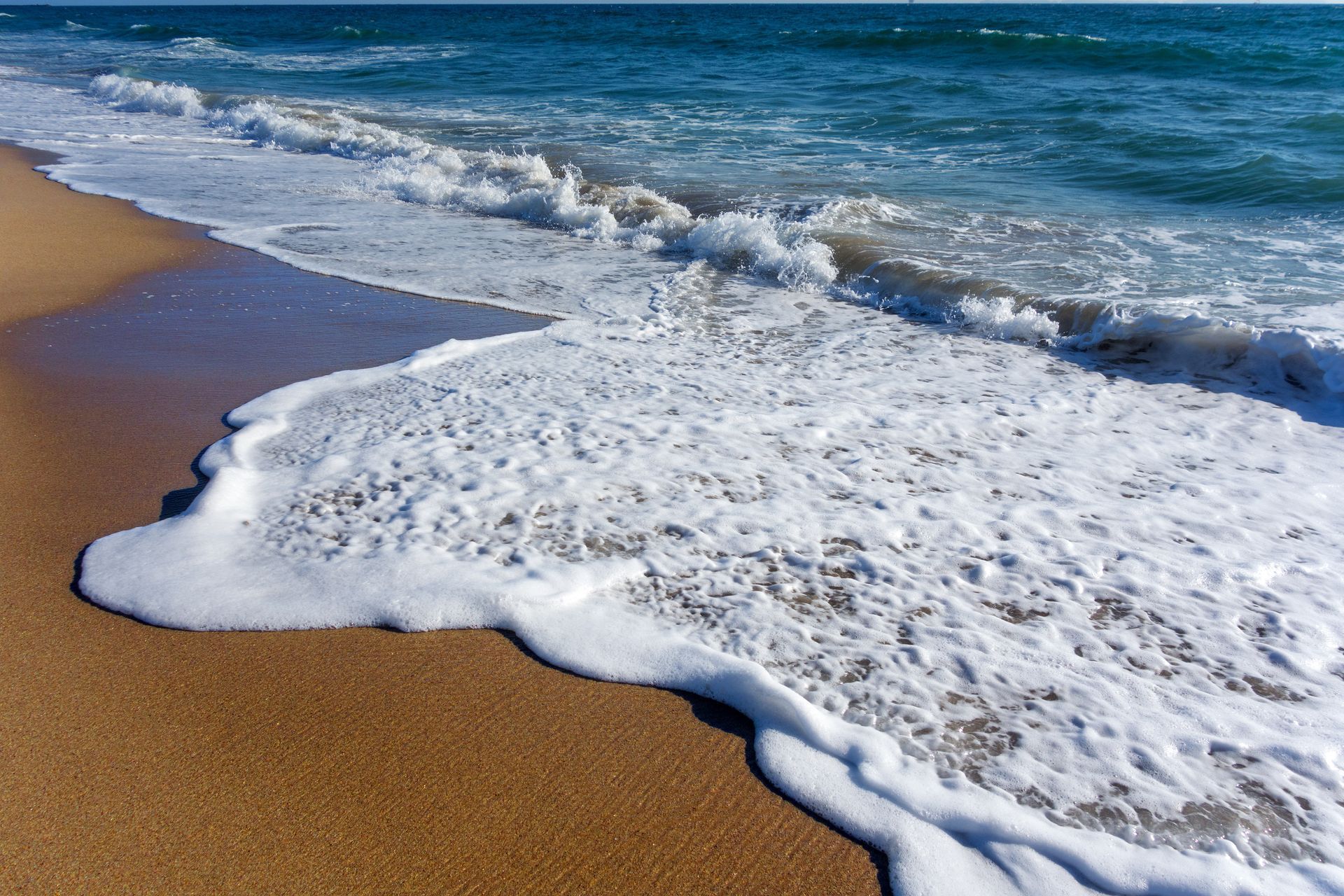 White sea foam washes over golden sand on a sunny beach, with turquoise ocean waves in the background.