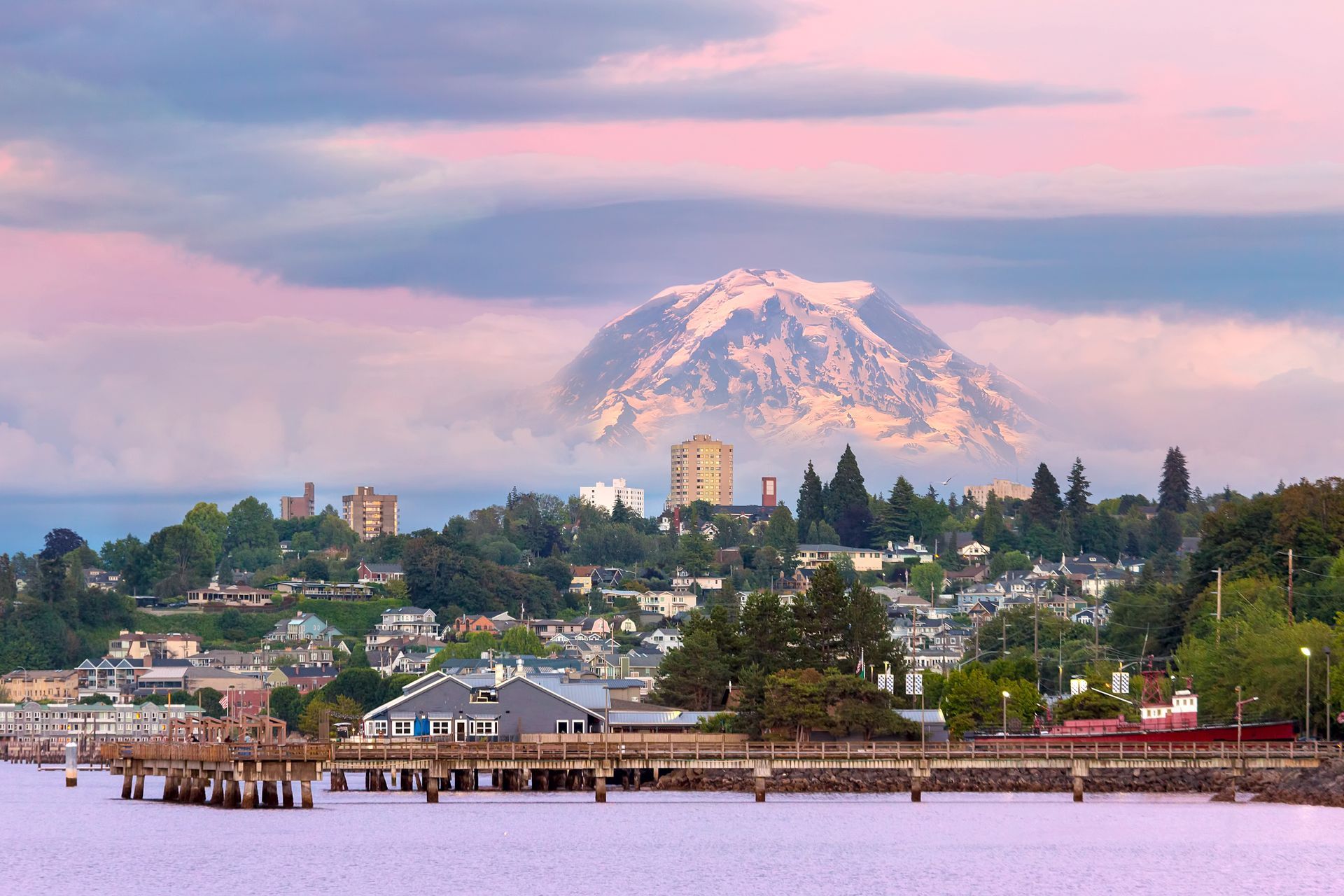 Snow-capped mountain above a waterfront town at sunset, with pink clouds and a pier in the foreground