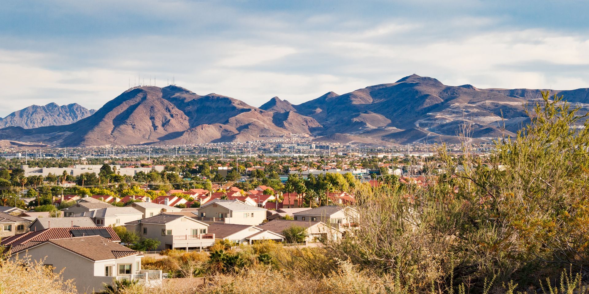 A sprawling suburban neighborhood sits in the foreground with a vast, arid mountain range under a cloudy sky behind it.