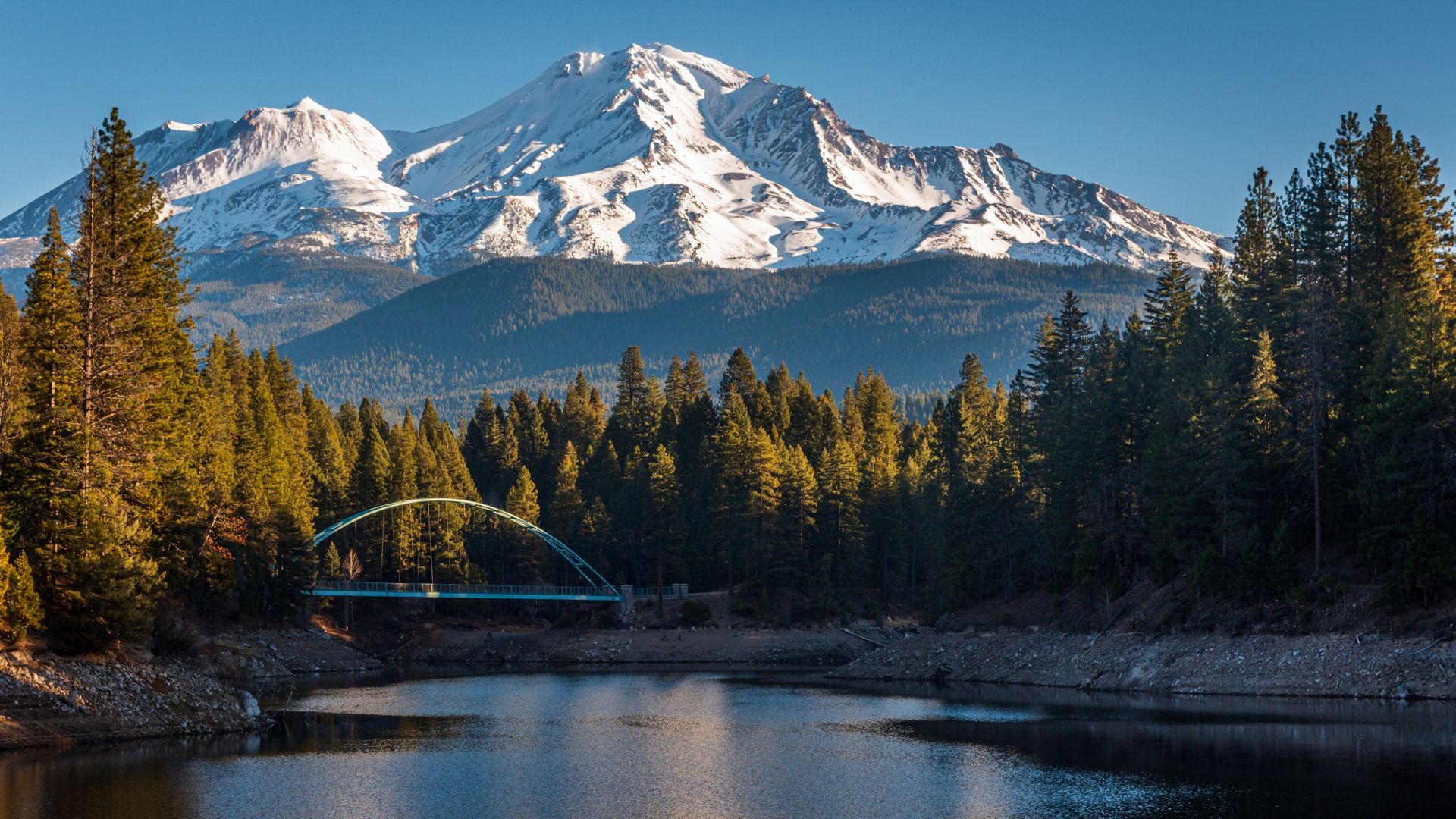 Snow-capped mountain rising above a lake and evergreen trees; a blue arched bridge crosses the water.