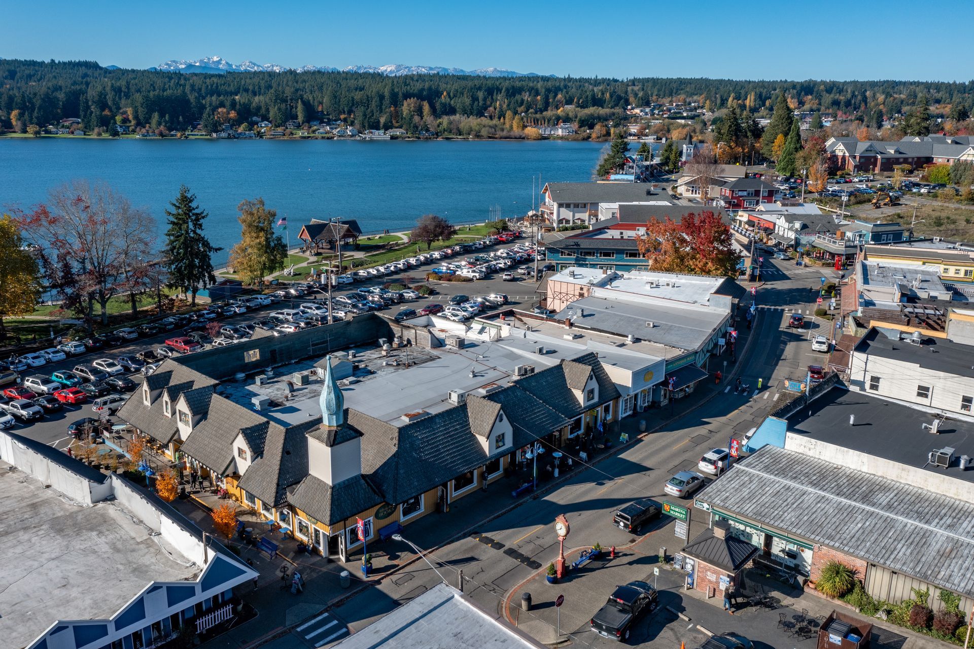 An aerial view of a shopping plaza with a clock tower, next to a lake with snow-capped mountains in the background.