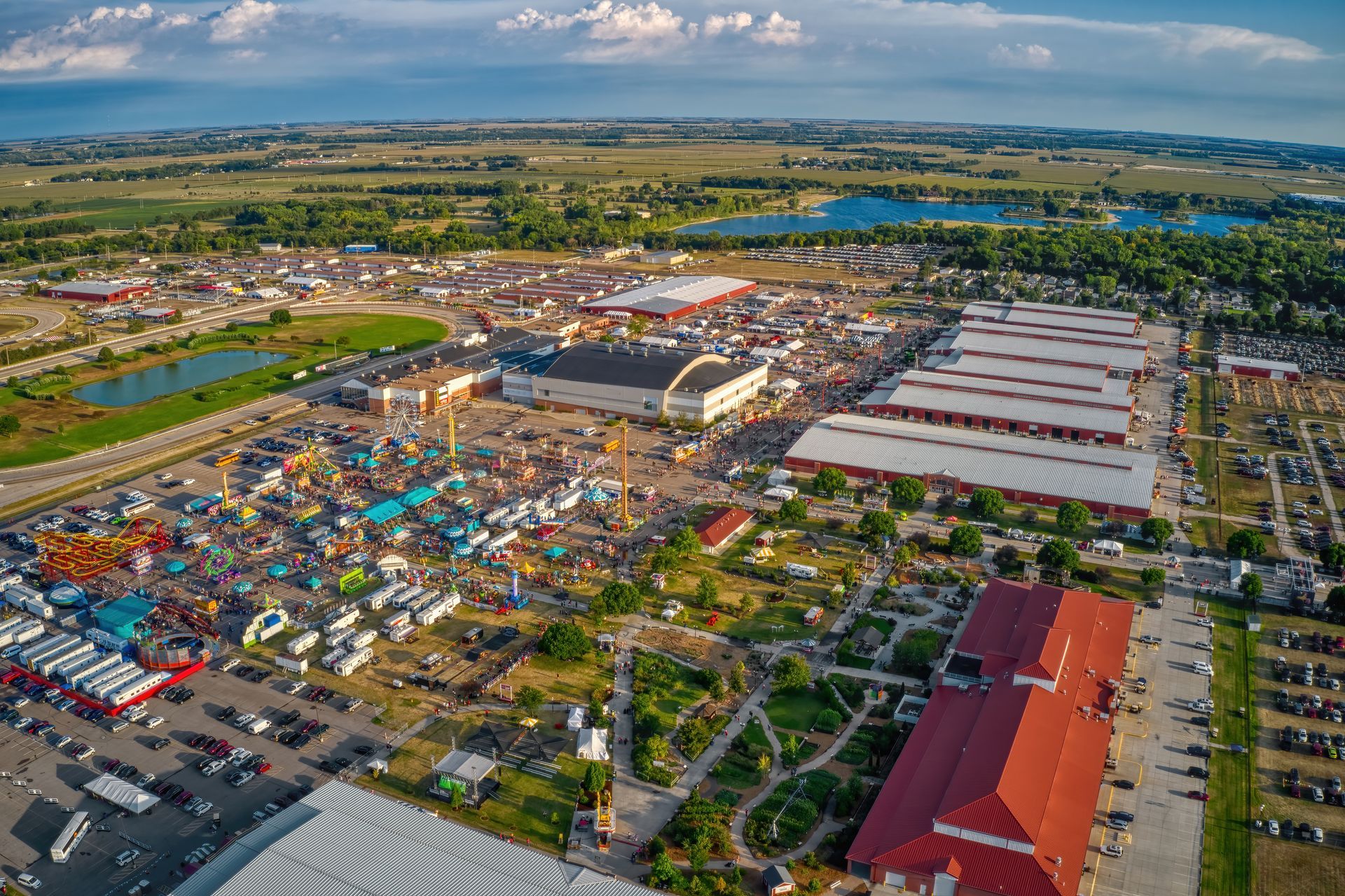 Aerial view of a  County Fair with rides, vendors, crowds, and buildings. Green fields and a lake are in the background.