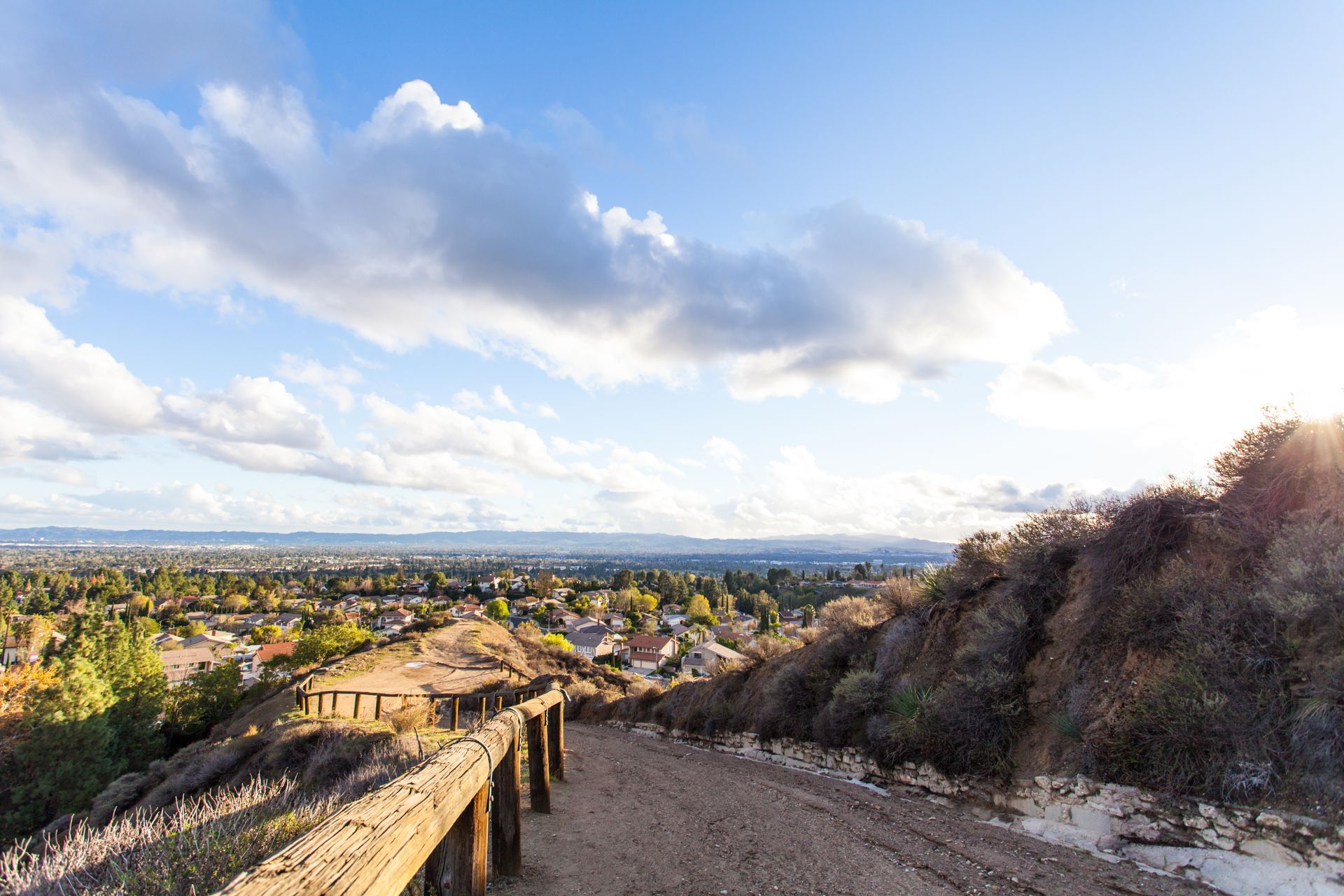 A dirt path winds down a hillside past a wooden railing, overlooking a Northridge, CA under a cloudy blue sky.