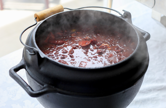 Panela fumegante de ensopado escuro com linguiça, feijão e legumes sobre uma toalha de mesa branca.